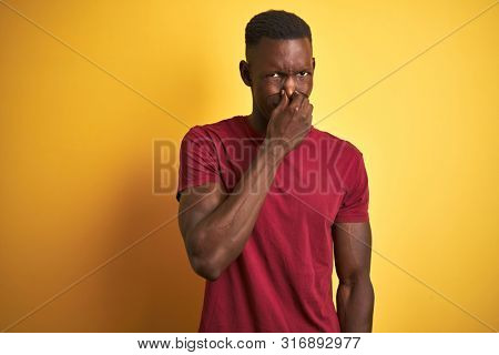 Young african american man wearing red t-shirt standing over isolated yellow background smelling something stinky and disgusting, intolerable smell, holding breath with fingers on nose. Bad smells