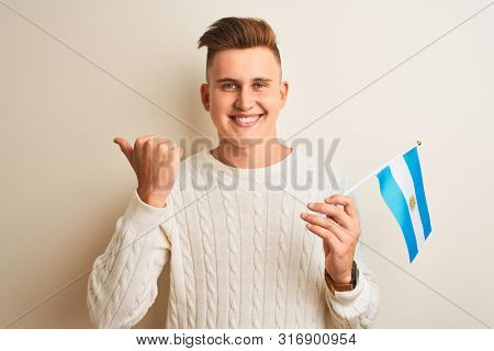 Young handsome man holding Argentinian Argentina flag over isolated white background pointing and showing with thumb up to the side with happy face smiling