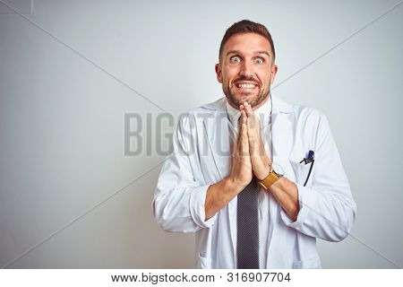 Young handsome doctor man wearing white profressional coat over isolated background praying with hands together asking for forgiveness smiling confident.