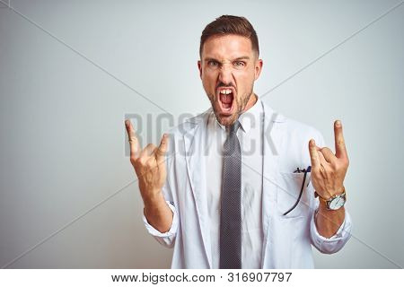 Young handsome doctor man wearing white profressional coat over isolated background shouting with crazy expression doing rock symbol with hands up. Music star. Heavy concept.