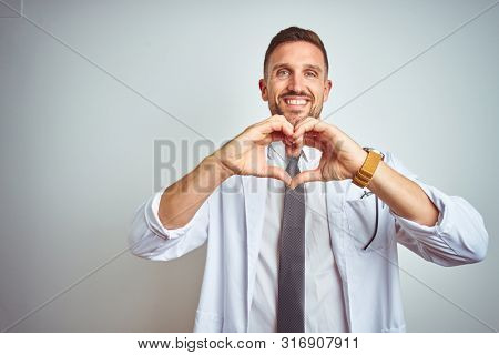 Young handsome doctor man wearing white profressional coat over isolated background smiling in love doing heart symbol shape with hands. Romantic concept.
