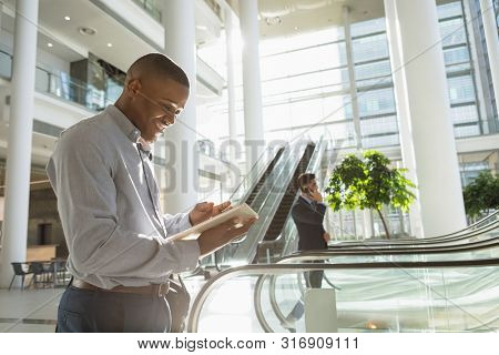 Side view of young African American businessman using tablet computer in the lobby of a modern office building. A young Caucasian businessman is talking on a phone and stepping on to an escalator in