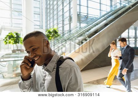 Close up side view of young African American businessman talking on smartphone by the escalators in sunlit modern business atrium. A young Caucasian businessman and businesswoman are walking together