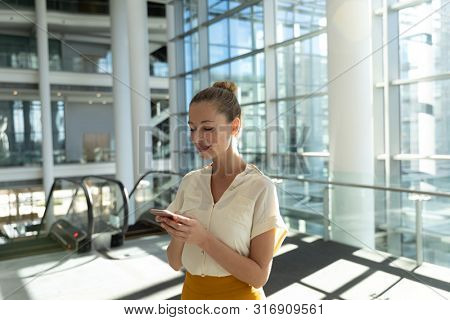 Side view of young Caucasian businesswoman using smartphone standing in front of escalators in the glass walled lobby of a modern business building. Modern corporate start up new business concept with