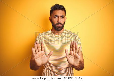 Young indian man wearing t-shirt standing over isolated yellow background Moving away hands palms showing refusal and denial with afraid and disgusting expression. Stop and forbidden.