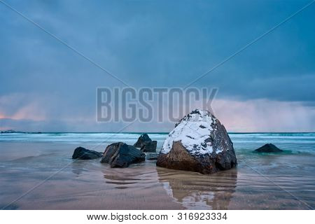 Rocks covered with snow on Norwegian sea beach in fjord in stormy weather with clouds. Skagsanden beach, Flakstad, Lofoten islands, Norway. Long exposure motion blur