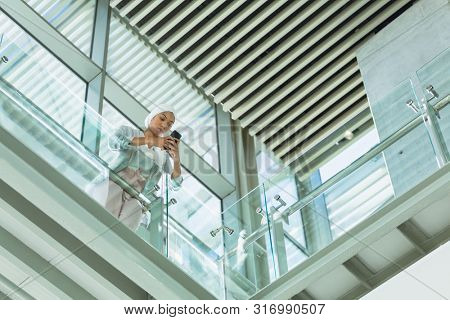 Low angle view of businesswoman in hijab leaning on railing and using mobile phone in corridor at modern office. Modern corporate start up new business concept with entrepreneur working hard