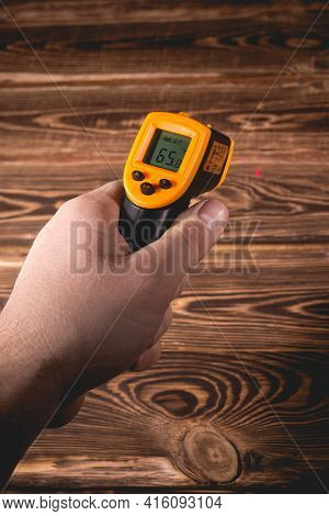 A Man Holds A Yellow-black Pyrometer On A Wooden Background. A Device For Non-contact Temperature Me