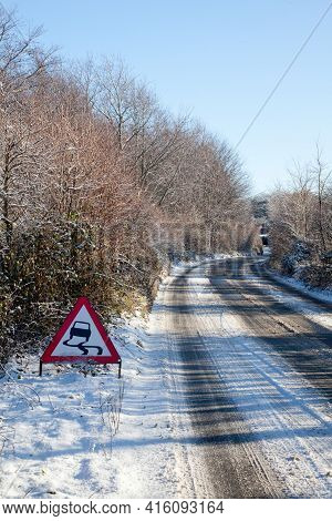 A Country Lane In The Snow On A Sunny Day With A Road Sign Warning Of Slippery Road Surface