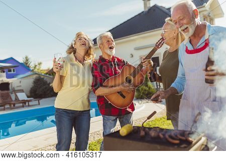 Group Of Cheerful Senior Friends Having A Poolside Backyard Barbecue Party, Gathered Around The Gril