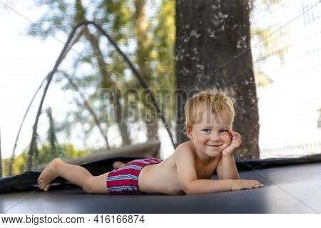 Portrait Of Cute Little Caucasian Funny Happy Blond Toddler Boy Lying Inside Big Black Trampoline At