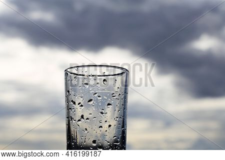Drinking Glass Covered With Water Drops On Background Of Dark Sky With Storm Clouds. Concept Of Rain