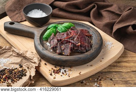 Slices Of Beef Jerky, Pepper, Salt, And Fresh Basil On A Beige Chopping Board. Jerky Meat In A Woode