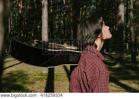 Portrait Of A Guy Walking Through The Woods With A Guitar In His Hands. Against The Background Of Tr