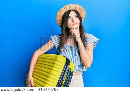 Young beautiful caucasian girl wearing summer dress and holding cabin bag serious face thinking about question with hand on chin, thoughtful about confusing idea 