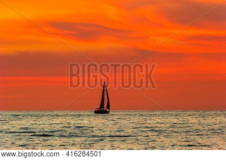 A Sailboat Is Sailing Along The Ocean As A Vivid Colorful Sunset Is In The Background