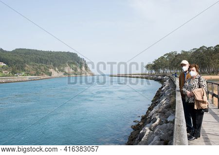 Aged Couple 70-80 Walking In Rodiles, Near The River And Puntal Forest In The Background, Asturias.