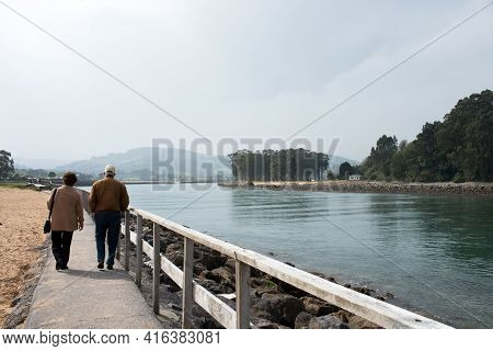 Aged Couple 70-80 Walking In Rodiles, Near The River And Puntal Forest In The Background, Asturias.