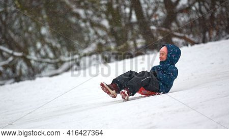 Moscow - 01.12.2021 - Cheerful Little Girl In Blue Down Jacket Is Rolling Down Hill On Red Sled. Win