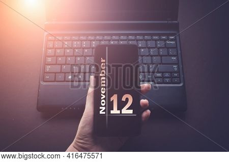 November 12nd. Day 12 Of Month, Calendar Date. Hand Holding Mobile Phone On Laptop Computer Dark Bac