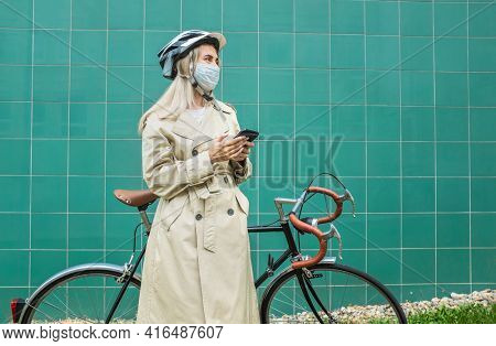 Woman in protective maskin, in helmet using her smartphone device. Female go to work on his sport retro bike. Girl standing on green background.