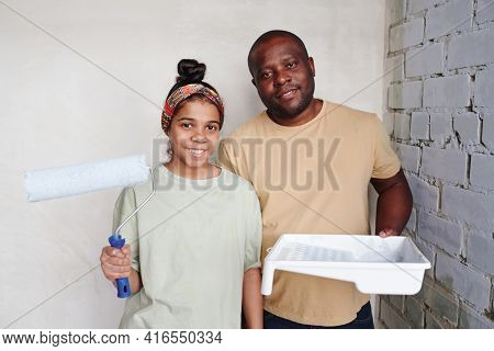 Happy cute teenage girl with paintroller and her smiling father with white square plastic container with paint standing in the corner