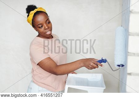Young African woman with paintroller standing in front of brick wall of living-room while painting it into white color during home renovation