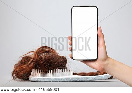 A White Comb With False Hair. White Background. A Female's Hand Holds A Smartphone With A White Scre