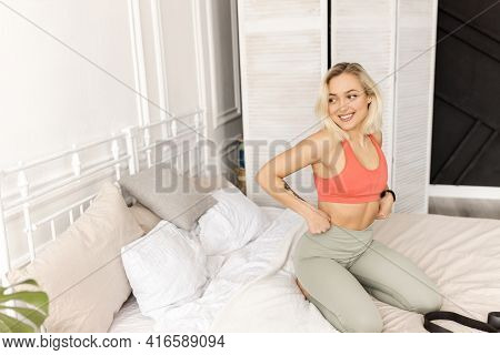 Young Blonde-hair Woman In Sportswear Is Sitting On A Bed, Getting Ready For Morning Workout. Sporti