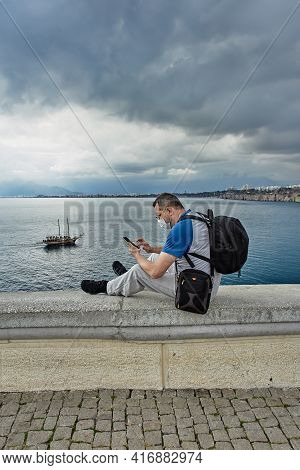An Independent Tourist Wearing Face Mask Looks Through Messages On Smartphone While Sitting On Guard