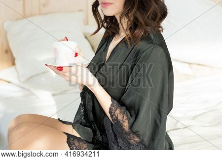 Girl In A Black Home Dressing Gown, Sitting On The Bed At Home And Drinking Coffee In The Morning