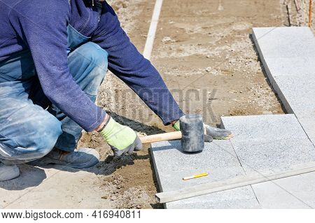 A Worker In A Blue Overalls, Half-crouched, Strikes The Granite Blocks With A Rubber Mallet, Assembl
