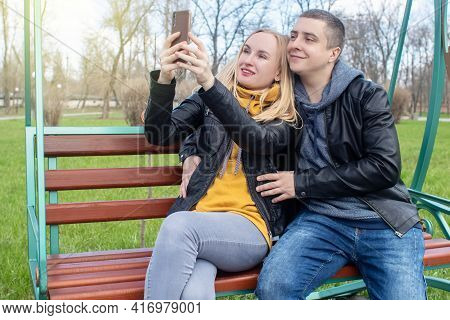 Husband And Wife Take A Selfie While Sitting On A Swing In The Park. Happy Couple Taking Pictures Of