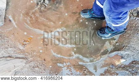 Close-up Of A Child's Feet In Rubber Boots, A Boy Child Happily Playing With A Shovel In A Muddy Pud