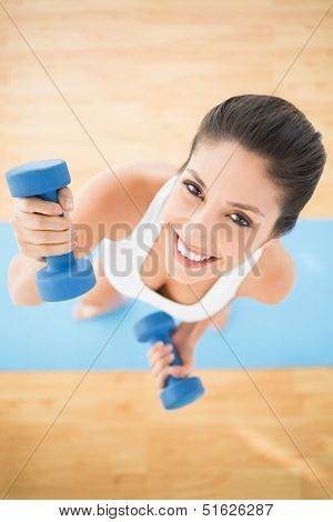 Happy woman exercising with dumbbells on blue exercise mat smiling at camera at home on wooden floor