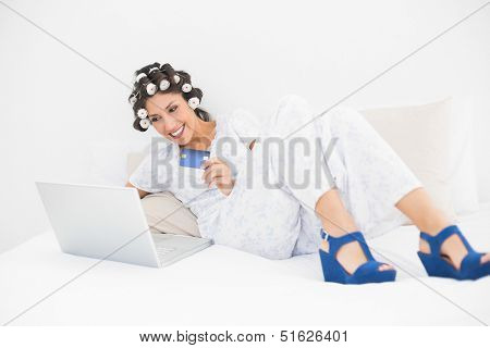 Brunette in hair rollers and wedge shoes using her laptop for shopping online in bedroom at home