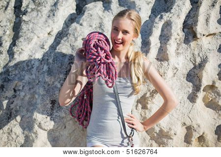 Smiling woman holding climbing equipment leaning at rock looking at camera