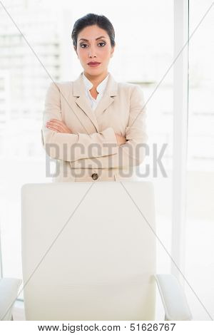 Serious businesswoman standing behind her chair in her office