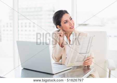 Smiling businesswoman holding newspaper while working on laptop looking away at the office