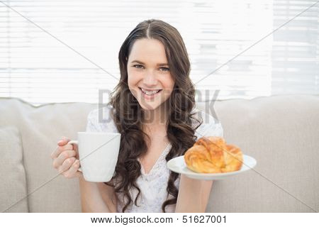 Smiling young woman in pajamas having breakfast while sitting on cosy sofa