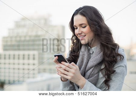 Cheerful pretty brunette in winter clothes sending a text on her smartphone outside on a cloudy day