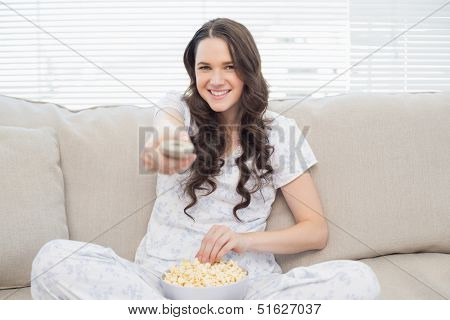 Cheerful woman in pajamas having popcorn while watching tv sitting on cosy sofa