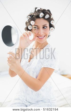 Pretty brunette in hair rollers looking in hand mirror and using eyelash curler in bedroom at home