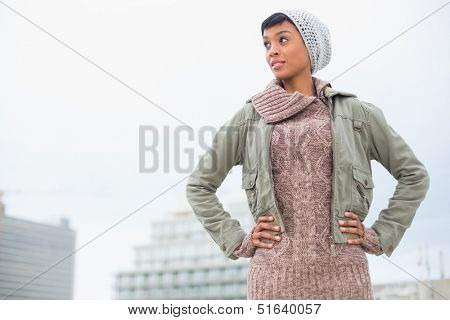 Thoughtful young model in winter clothes posing hands on the hips outside on a cloudy day