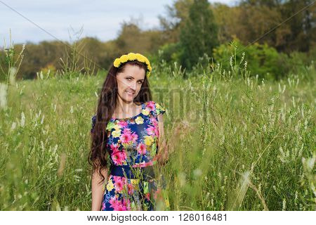 Beautiful woman with long hair and a wreath of flowers on his head walking in a field among the tall grass in warm summer evening