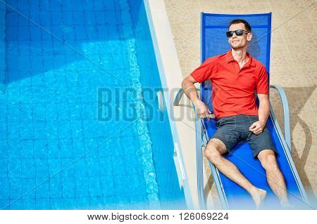 young man relaxing on the chaise-longue near the pool. man wearing in red polo shirt and sunglass
