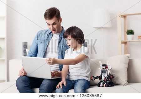 Having leisure time. Cheerful surprised positive father and his son sitting on the sofa and expressing joy while using laptop