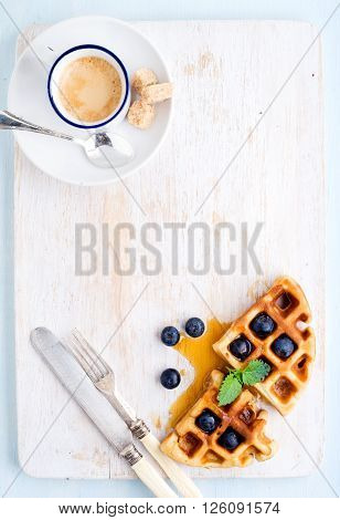 Espresso coffee cup and soft belgian waffles with fresh blueberries and marple syrup on white painted wooden board over light blue background. Top view, copy space