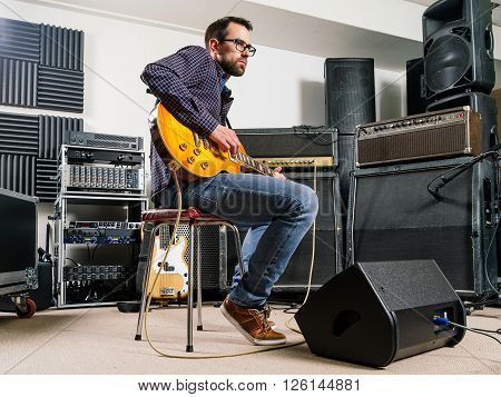 Photo of a man in his late 20's sitting in a recording studio playing his electric guitar.