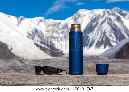 Blue Travel Thermos with Opened Cup and Sunglasses on Wooden Table High Majestic Himalaya Mountain Range with Snow and Ice on Background